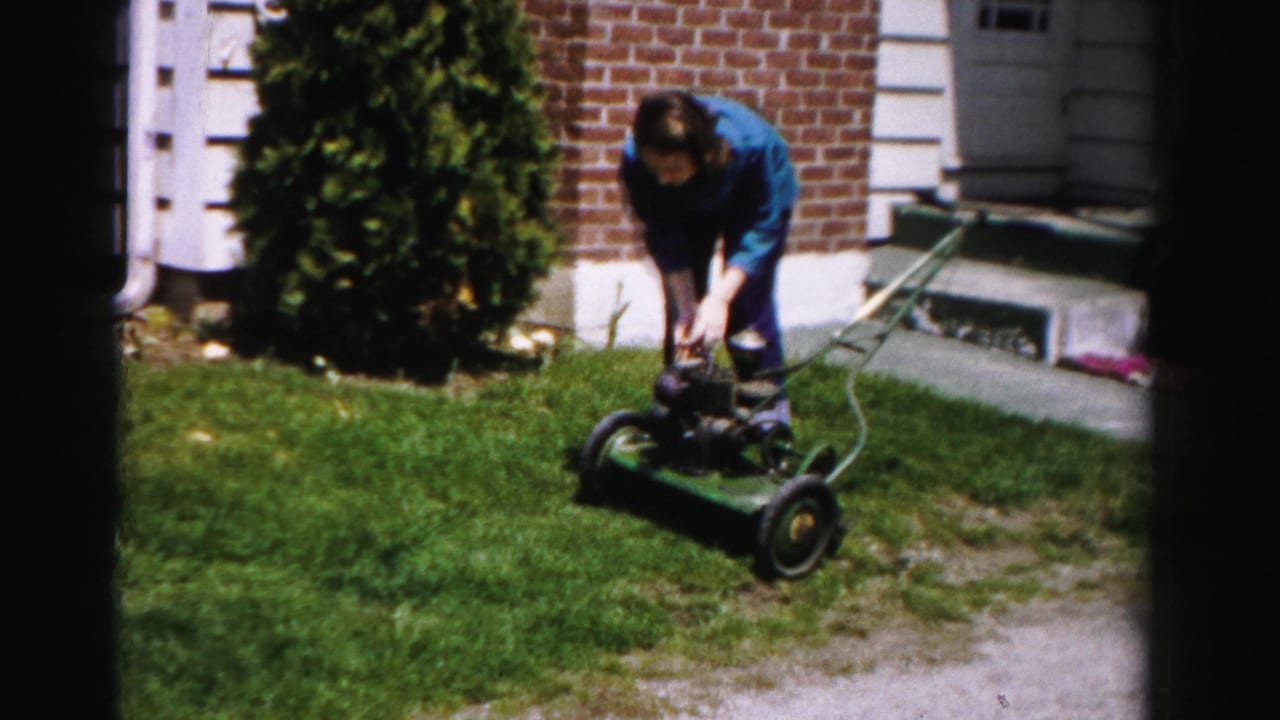 Woman leans forward pushing a green rotary mower along a suburban front lawn; brick and white-trim house with front entrance steps visible; Albany, New York, mid-1950s.