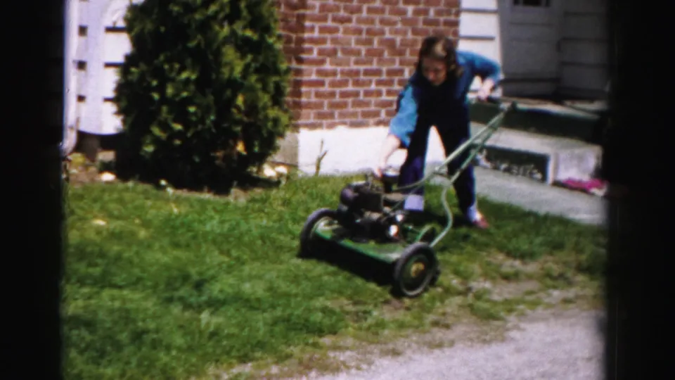 Woman bending over a green rotary push mower on a suburban front lawn; brick house foundation, white-painted trim, and evergreen shrub visible; mid-1950s home movie.
