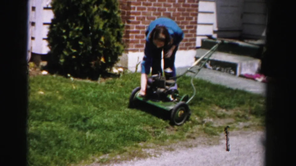 Woman pushes green rotary mower near brick house front steps; film sprocket-hole artifact at lower right; mid-1950s 8mm home movie, Albany, New York.