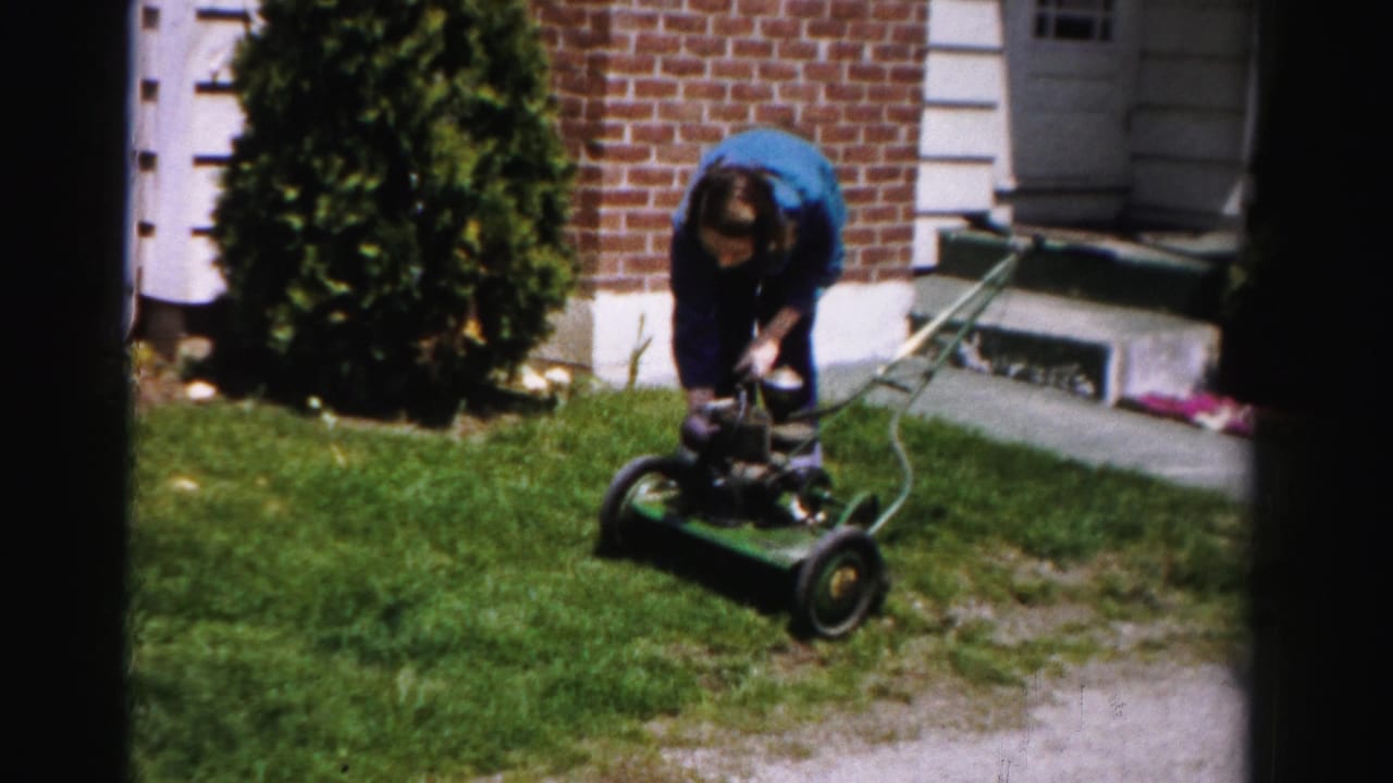 Person attempting to start a green push lawn mower in front of a brick suburban house with white trim, mid-1950s Albany, New York area, 8mm home movie.