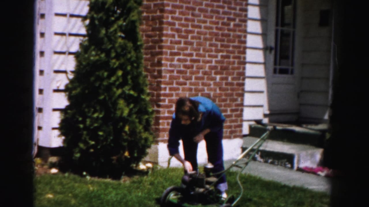 Person in blue top leaning toward a green push mower beside columnar arborvitae, brick house with white-trimmed steps in background.