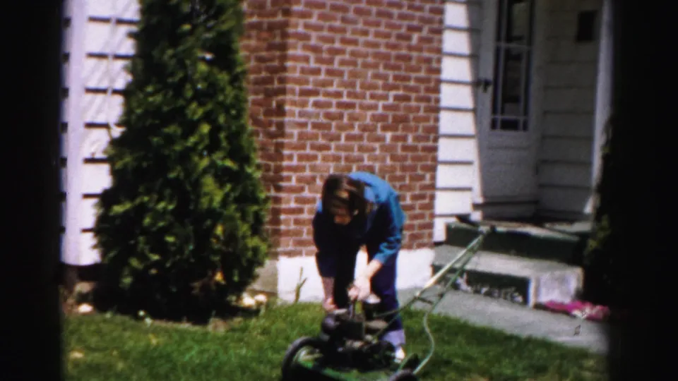 Person crouching to work on a green push mower engine in a mid-1950s suburban yard, brick house facade, small pink flowers near steps visible.