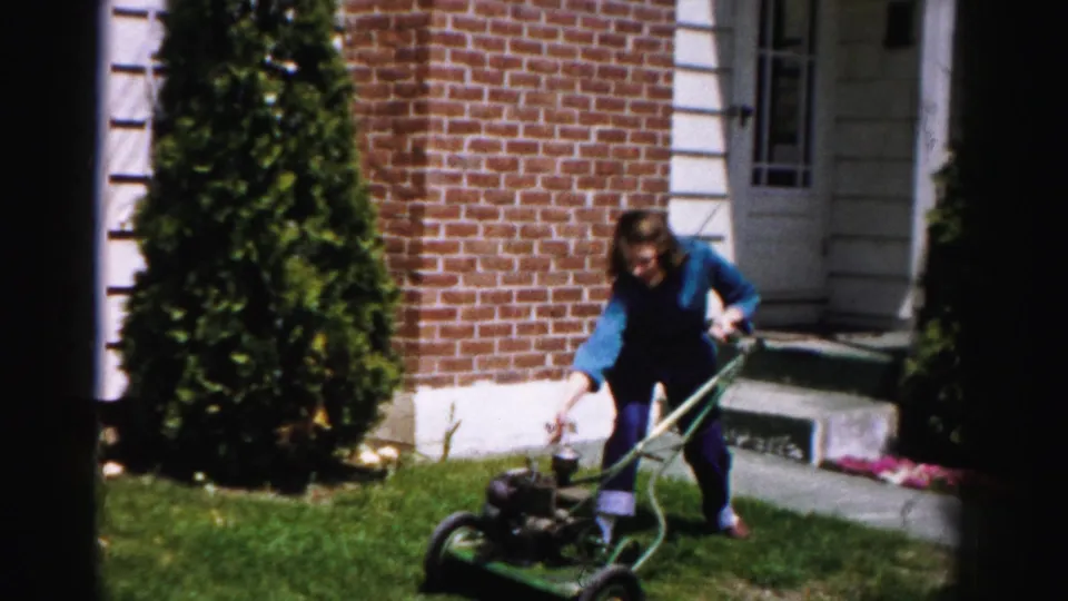 Person gripping the handle of a green push mower in a mid-1950s suburban yard, brick house with white foundation trim and arborvitae shrubs visible.