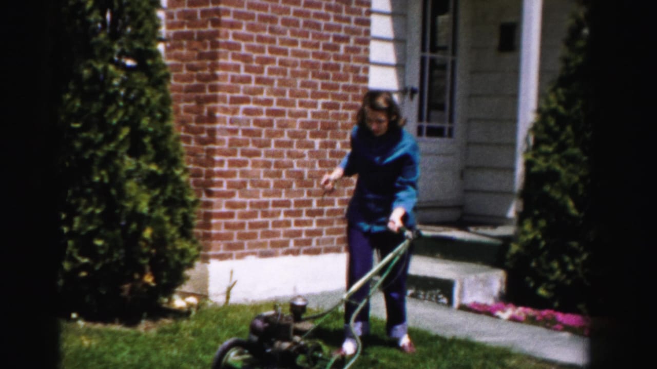 Woman in blue outfit stands beside a vintage green push lawn mower on a lawn in front of a brick house with tall arborvitae shrubs.