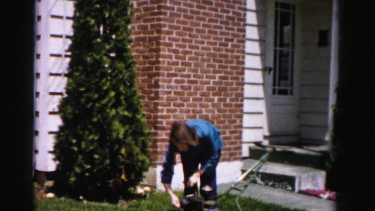 Woman crouching over the engine of a vintage push lawn mower in front of a brick-and-white-trim house, circa mid-1950s, Albany, New York area.