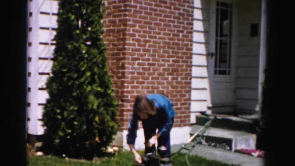 Woman crouching over the engine of a vintage push lawn mower in front of a brick-and-white-trim house, circa mid-1950s, Albany, New York area.