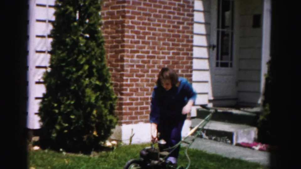 Woman leaning over a vintage gas push mower attempting to start it, front yard of brick house with white quoined trim, circa mid-1950s, Albany, New York area.