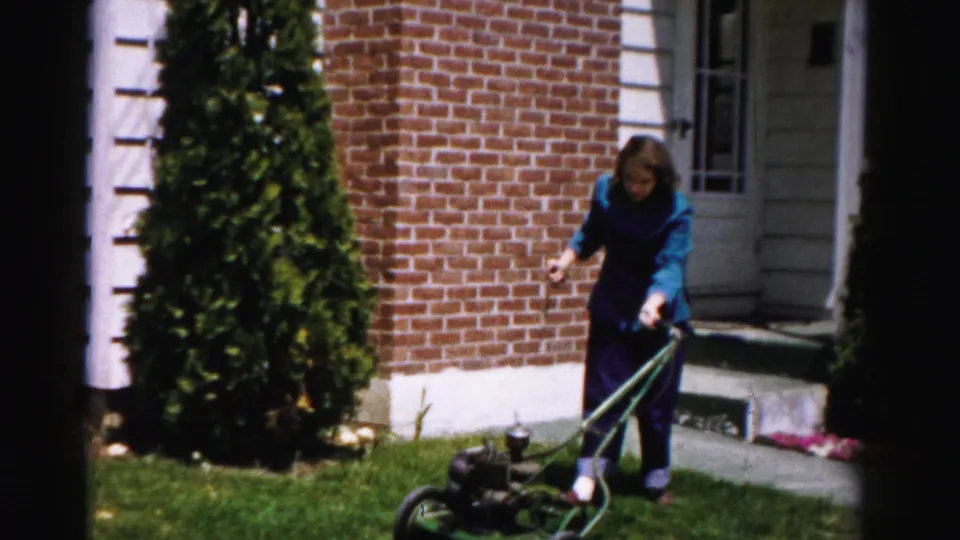 Woman standing at the handle of a vintage push lawn mower in the front yard of a brick house with white quoined trim, circa mid-1950s, Albany, New York area.