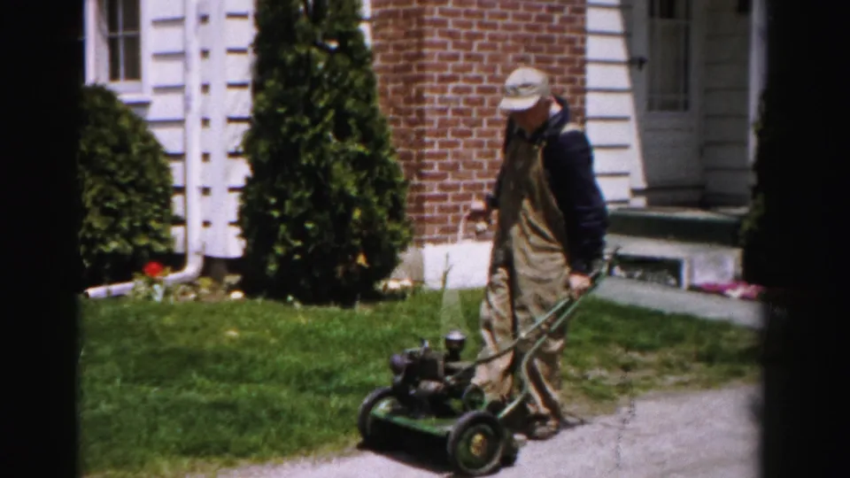 Man pausing beside a gasoline push mower in a tidy suburban front yard, Albany, New York, circa mid-1950s, 8mm home movie.