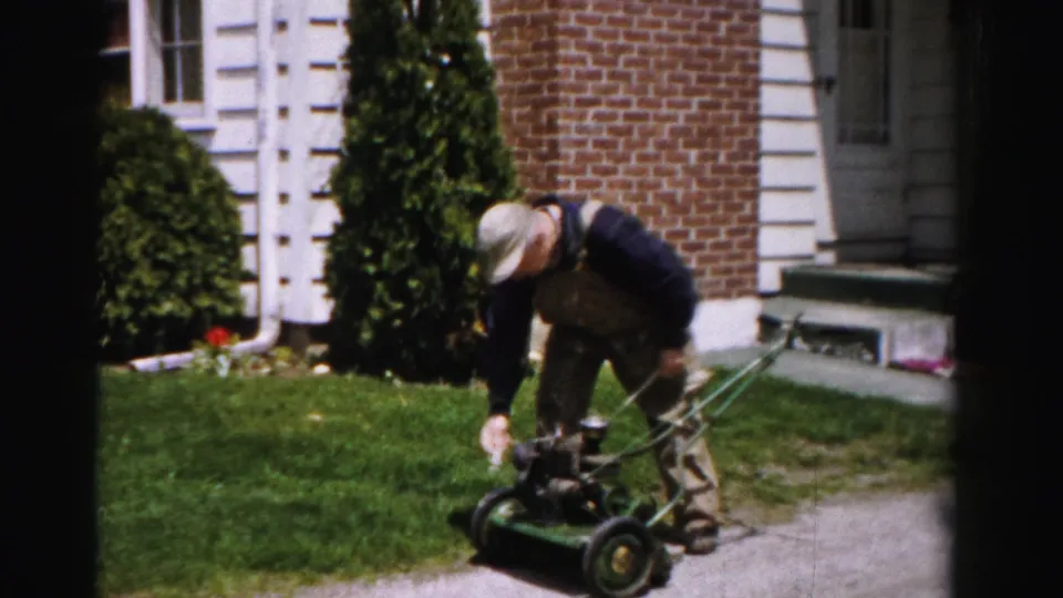 Man leaning into a gasoline push mower attempting to start it in a suburban yard, Albany, New York, circa mid-1950s, 8mm film.