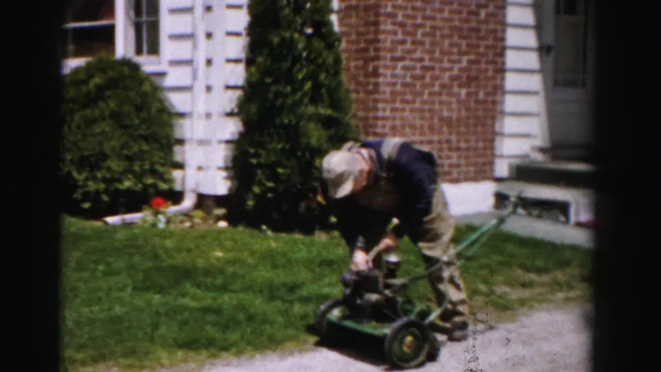 Man working to start a gasoline push mower near the corner of a brick suburban house, Albany, New York, circa mid-1950s, 8mm film.