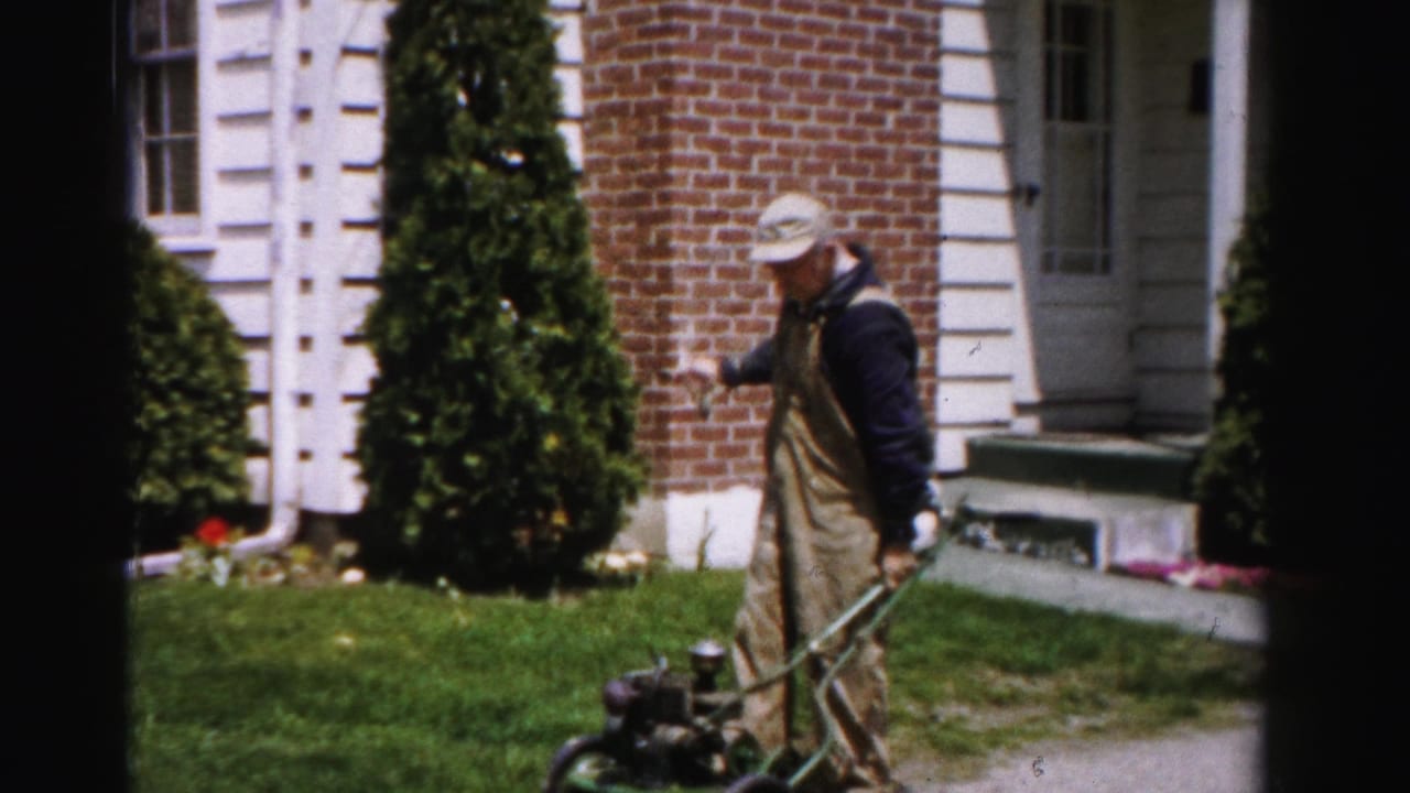 A man in tan bib overalls and a light cap stands upright beside a green push lawn mower in front of a brick and white-trimmed suburban house, circa mid-1950s Albany, New York.