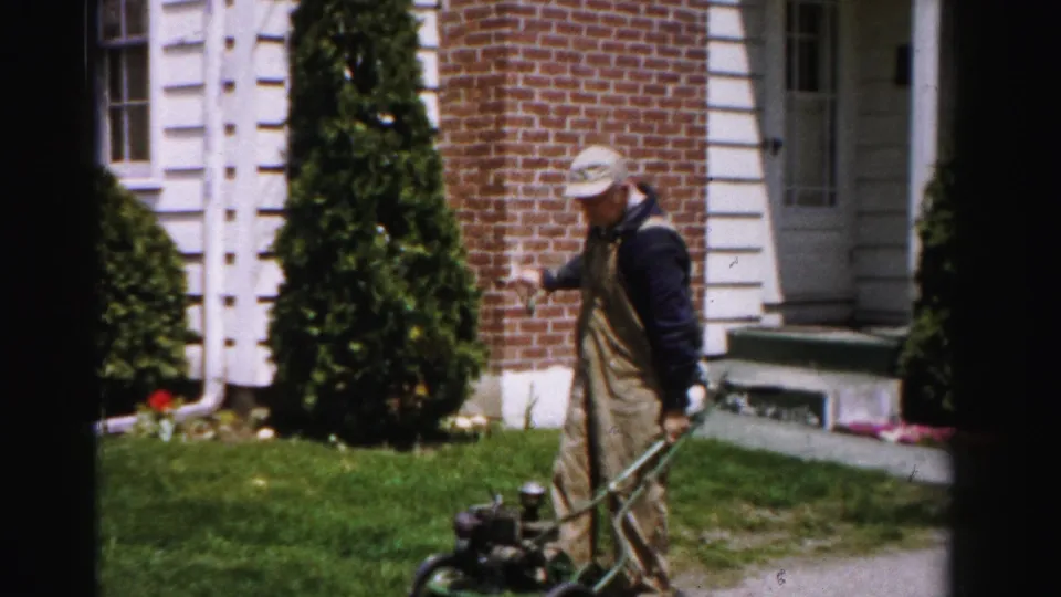 A man in tan bib overalls and a light cap stands upright beside a green push lawn mower in front of a brick and white-trimmed suburban house, circa mid-1950s Albany, New York.