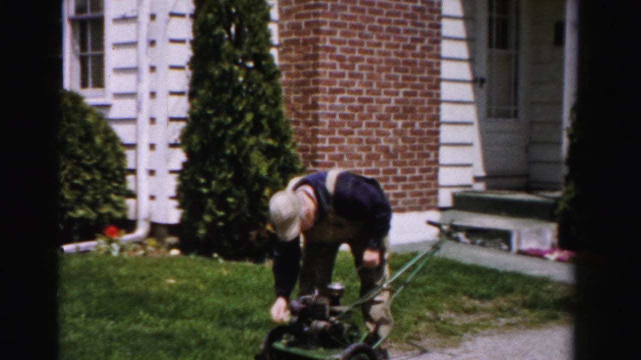 Man bends over a green gas push mower on a suburban front lawn, working on the starter mechanism.