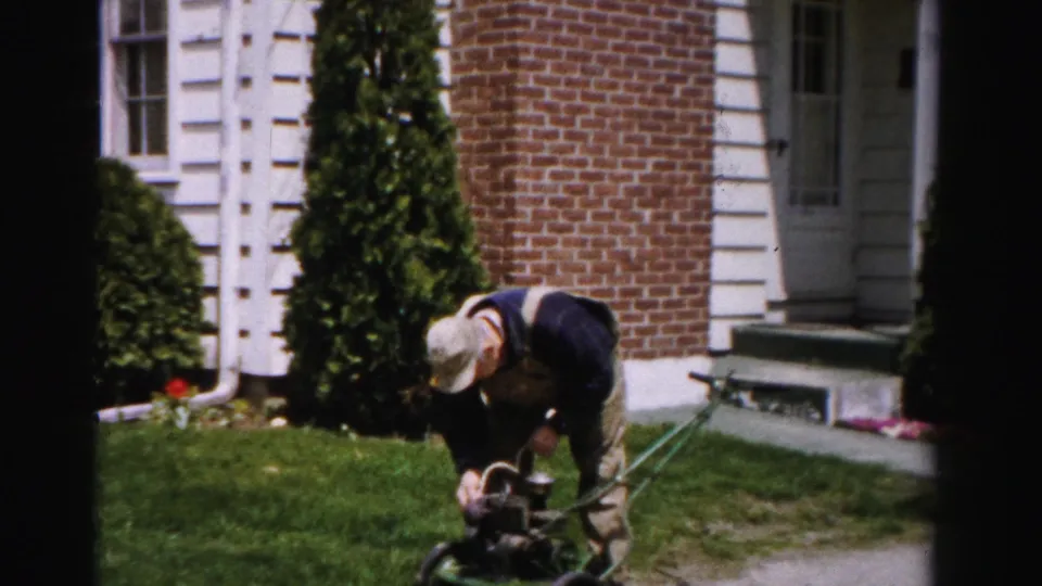 A man in bib overalls crouches deeply over a green gas push mower in a suburban front yard, brick house and white clapboard siding visible behind him, circa mid-1950s Albany, New York.