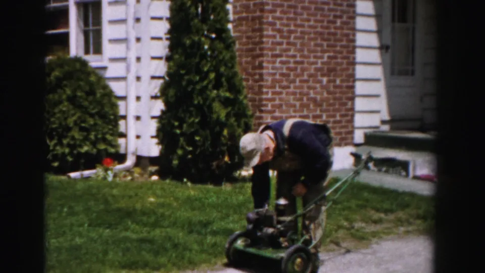 A man in bib overalls and a light cap leans over a green gas push mower on a front lawn, with a brick house and trimmed evergreen shrubs visible behind him, circa mid-1950s Albany, New York.