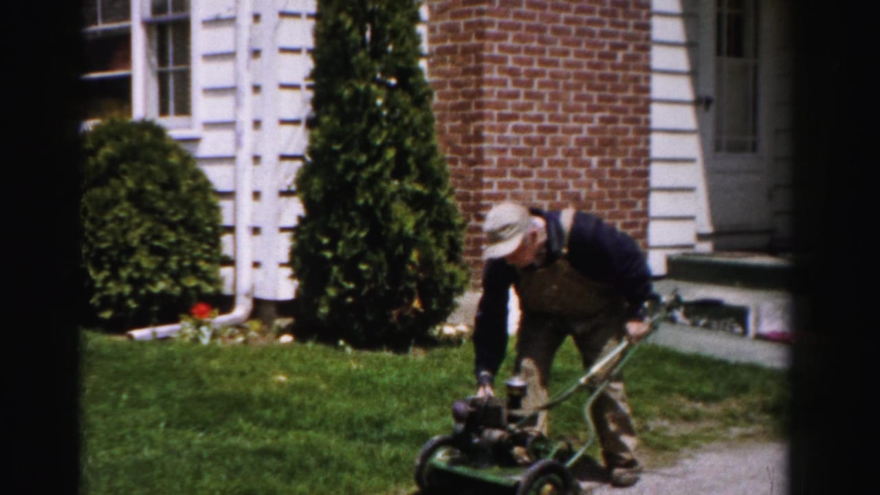Man in bib overalls bending over a green pull-start push mower in front of a brick house with white trim, Albany, New York, c.1955.