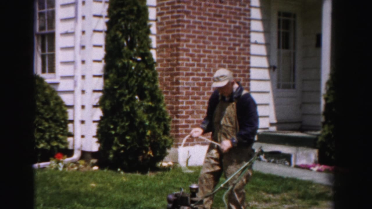 Man in overalls standing upright pulling the starter cord on a green push mower.