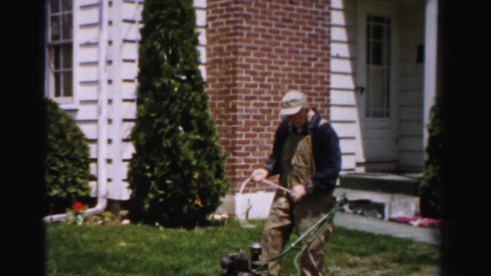 Man in bib overalls pulling the starter cord on a green push mower in front of a brick house, c.1955 Albany, New York.
