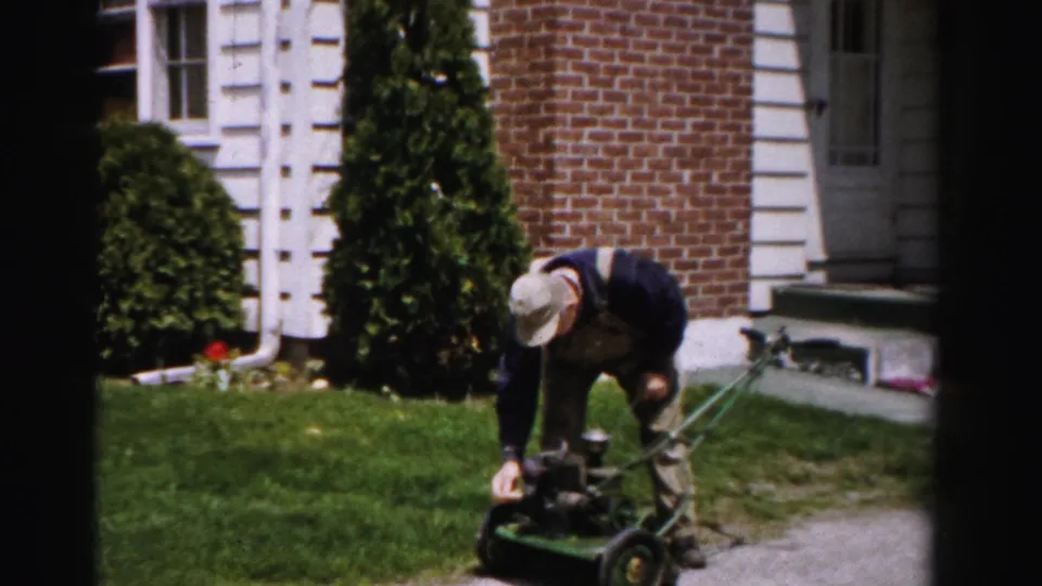 Man in cap and bib overalls crouched over a green push mower with a brick suburban house behind him, c.1955 Albany, New York.