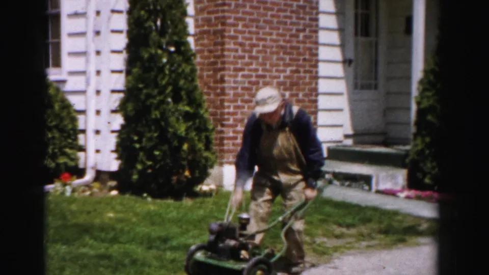 Man in bib overalls attempting another starter-cord pull on a green push mower in front of a brick house with white trim, c.1955 Albany, New York.