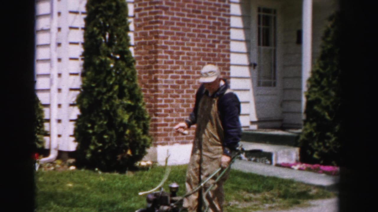 Man in bib overalls and bucket hat stands on a suburban lawn holding a reel-type push mower, brick house with arborvitae shrubs behind him.