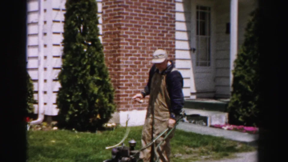 Man in bib overalls and cap stands on the front lawn of a brick suburban home holding a reel push mower; Albany, New York area, circa mid-1950s.