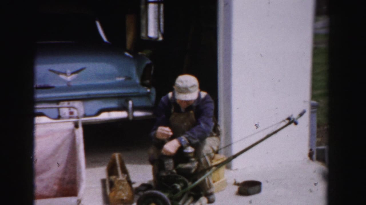 Man in bib overalls kneels on a driveway repairing a reel-type push mower; a blue late-1950s automobile with chrome V-shaped emblem is parked in the garage, Albany, New York area, circa mid-1950s.