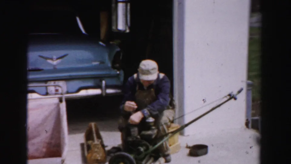 Man in bib overalls kneels on a driveway repairing a reel-type push mower; a blue late-1950s automobile with chrome V-shaped emblem is parked in the garage, Albany, New York area, circa mid-1950s.