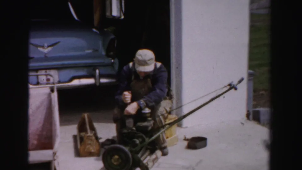 Man in bucket hat and bib overalls works on a disassembled reel mower on a suburban driveway; blue late-1950s automobile with chrome hood emblem parked behind him, circa mid-1950s.