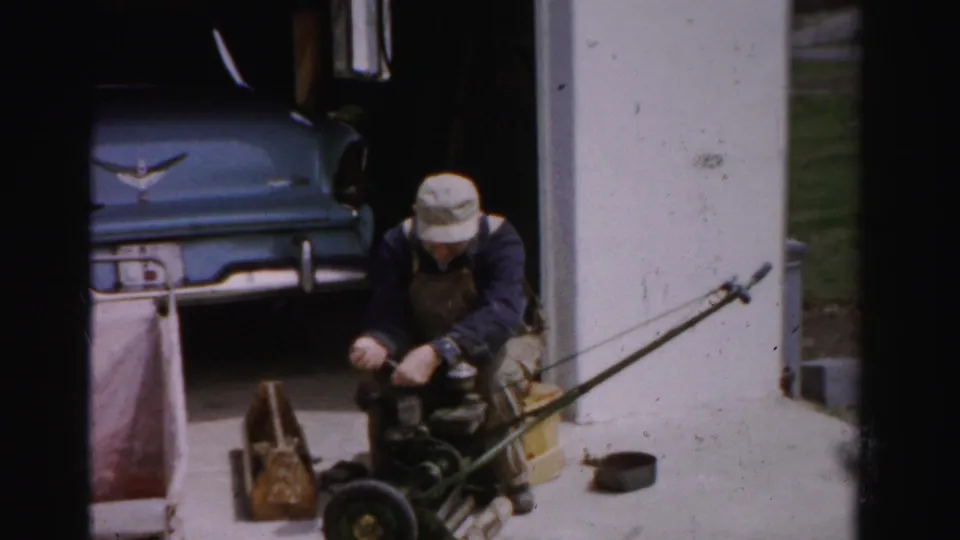 Man in bib overalls and cap kneels over disassembled lawn mower parts near a garage; blue late-1950s car with chrome emblem parked inside, Albany, New York area, circa mid-1950s.