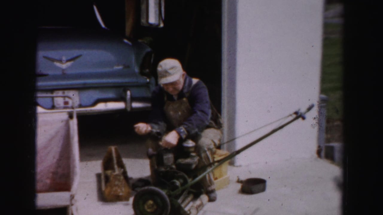 Mid-1950s man in bib overalls servicing a push reel mower outside a garage, blue American automobile with chrome ornament behind, Albany NY area.