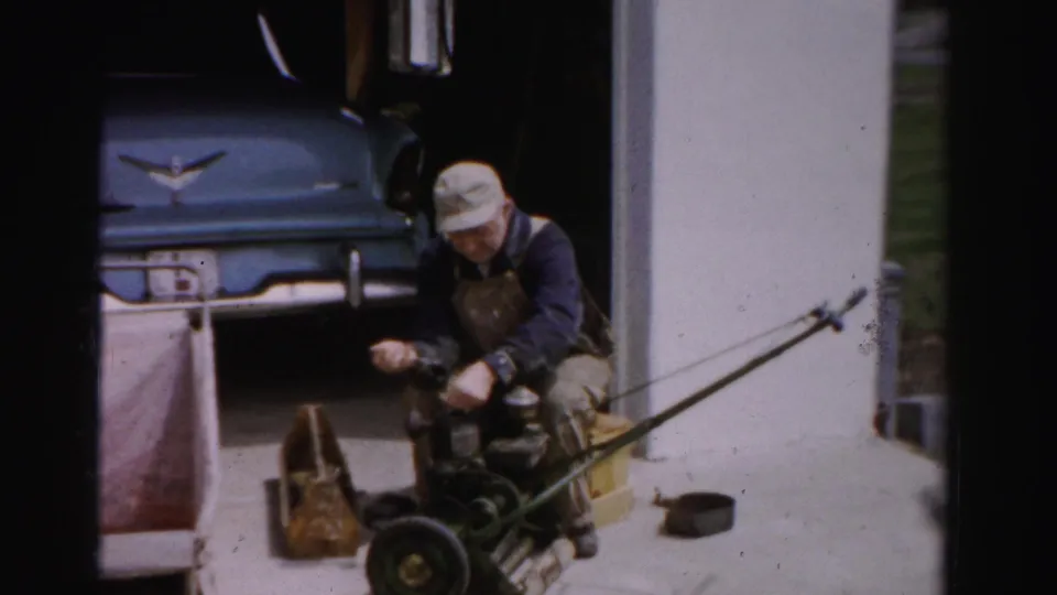 Mid-1950s man in bib overalls servicing a push reel mower outside a garage, blue American automobile with chrome ornament behind, Albany NY area.
