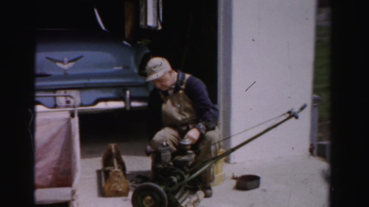 Man in bib overalls adjusting a push reel mower outside a garage, blue 1950s American car with chrome ornament behind, green lawn visible, Albany NY.