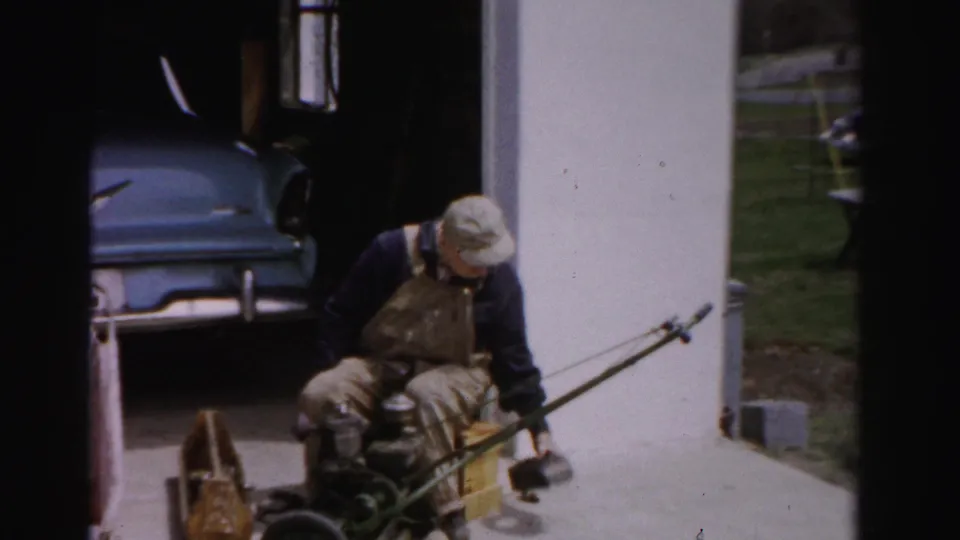 Man in bib overalls bent close over a push reel mower on a concrete driveway, blue 1950s American car in garage opening behind him, Albany NY.