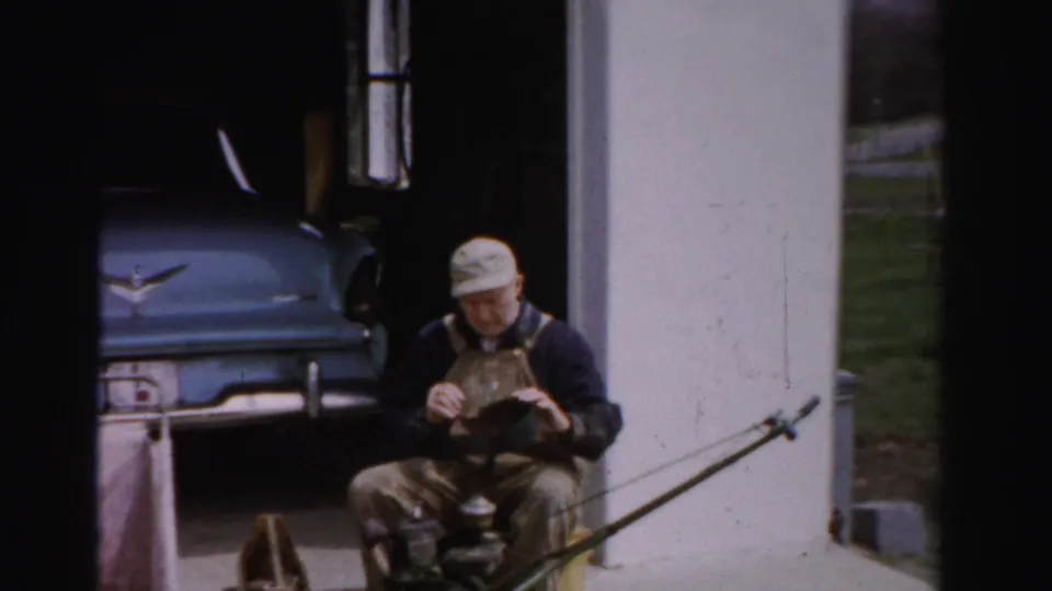 Man in bib overalls holding and inspecting a mower component outside a garage, blue 1950s American automobile and green lawn behind him, Albany NY area.