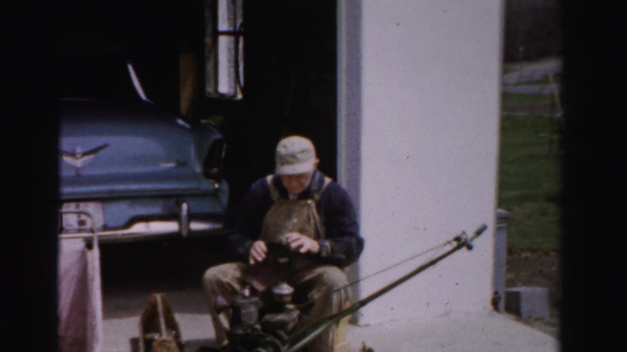Man in cap and overalls seated in a driveway, working on a small object, with a blue vintage American car behind him.