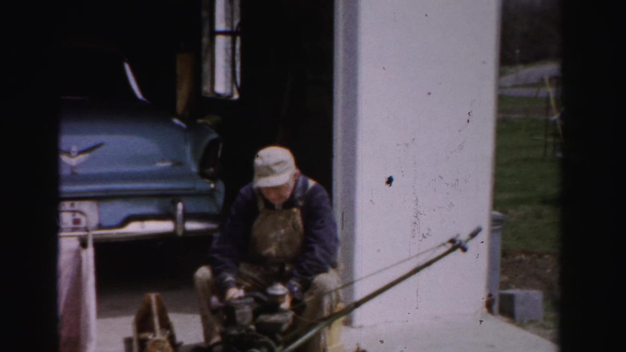 Continued shot of a man in work attire seated before a mid-1950s blue automobile in a driveway, tinkering with a small device; Albany, New York area, circa 1955-1956.