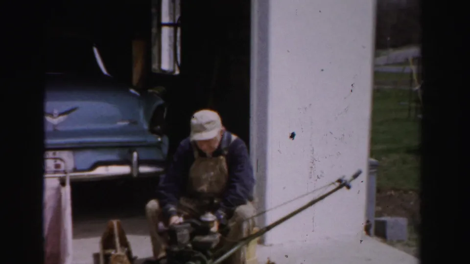 Continued shot of a man in work attire seated before a mid-1950s blue automobile in a driveway, tinkering with a small device; Albany, New York area, circa 1955-1956.