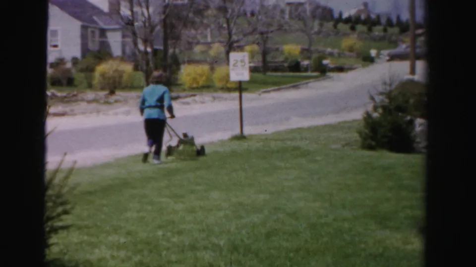 Woman in blue jacket and dark pants mowing a lawn in a mid-1950s suburban neighborhood; a 25 mph road sign, leafing trees, and yellow forsythia mark the early-spring setting; Albany, New York area.