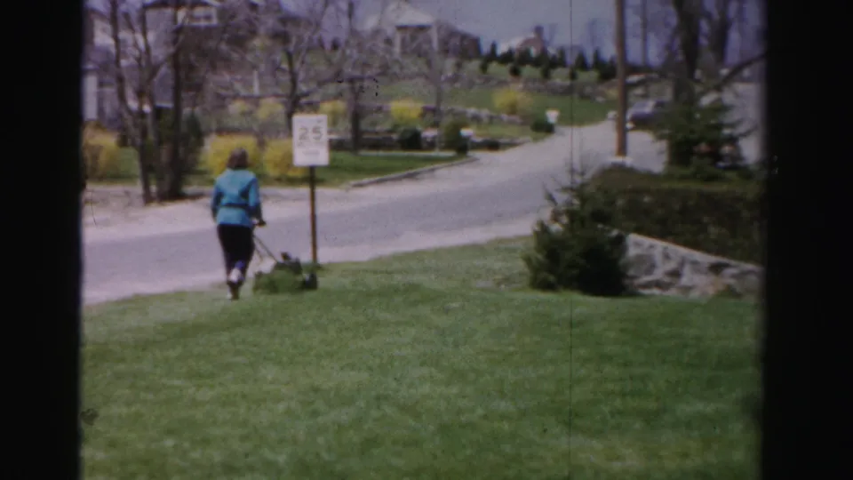 Woman mowing a lawn in an early-spring 1950s residential neighborhood; a 25 mph road sign, yellow forsythia, and neighboring homes are visible; Albany, New York area.