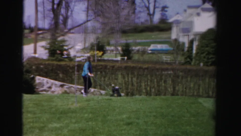 Wider view of a woman mowing alongside a low hedge in a mid-1950s Albany, New York residential yard; white house, bare deciduous trees, and evergreen shrubs in background.