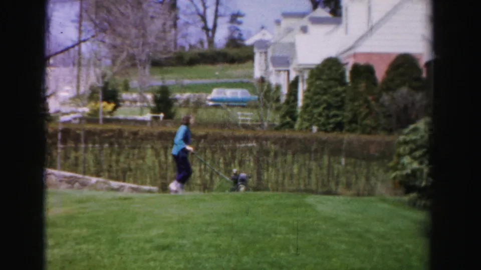 Woman continues mowing near a low hedge in a mid-1950s Albany, New York suburban yard; white house, bare trees, and evergreen conifers visible in background.