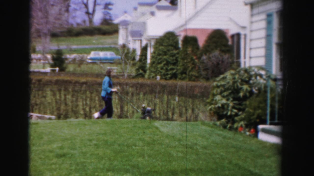 Young woman in blue jacket pushes a reel mower across a bright green lawn, white house with teal shutters behind her.