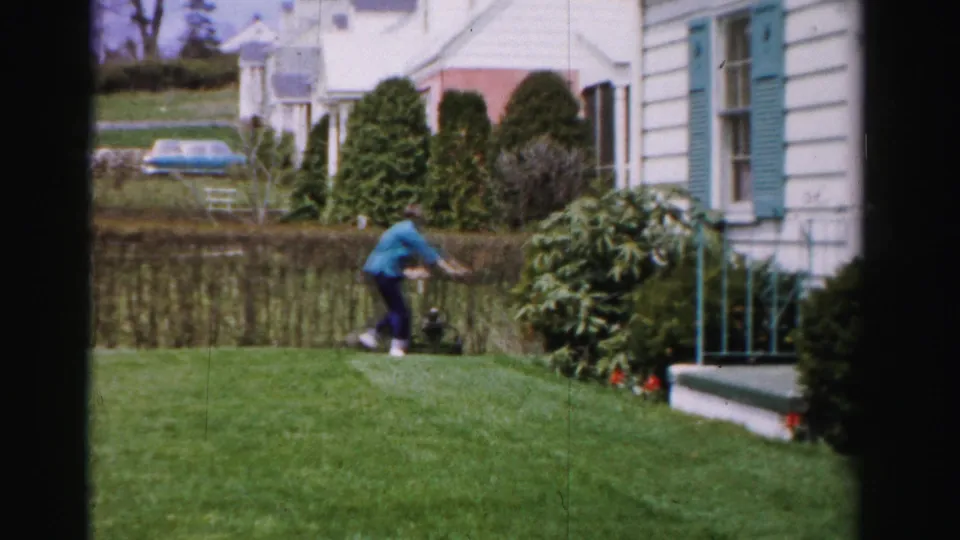 Woman pushing a reel mower near the front steps of a white suburban house, teal shutters and red flowering plants visible at base of steps, 1950s Albany New York.