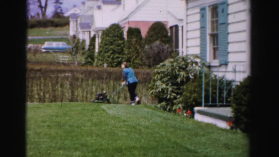 Woman mid-stride pushing a reel mower across a green suburban lawn, white clapboard house with teal shutters visible behind the hedge, 1950s Albany New York.