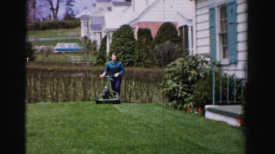 Woman pushing reel mower toward camera with face partially visible, white clapboard house with teal shutters and ornamental shrubs behind her, 1950s Albany New York.