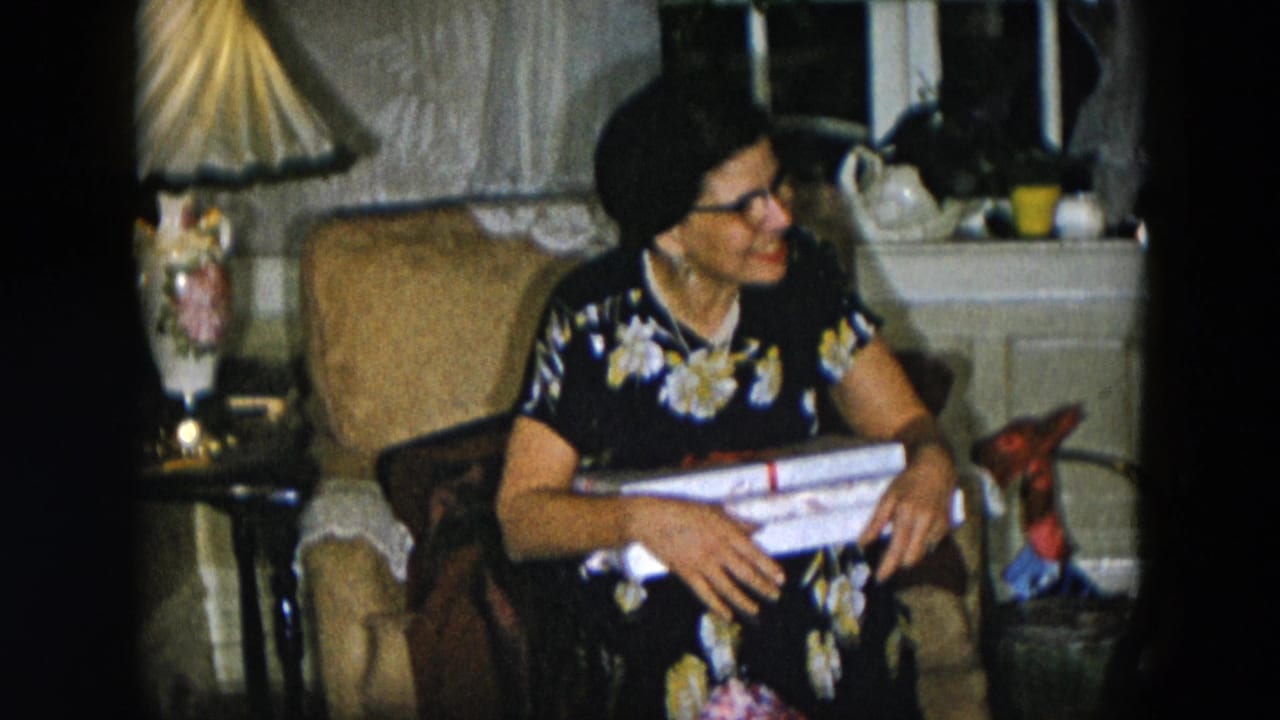Woman in 1950s floral housedress and cat-eye glasses seated in armchair holding wrapped presents, domestic living room interior, Hicksville New York, circa 1956.