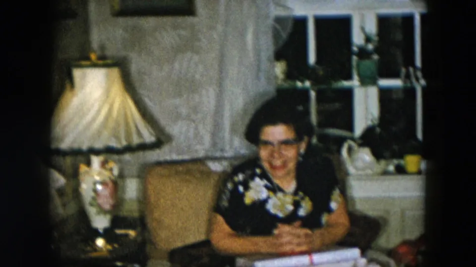 Woman in dark floral dress and cat-eye glasses facing camera with gifts on lap, large fabric table lamp behind her, 1950s domestic living room interior.