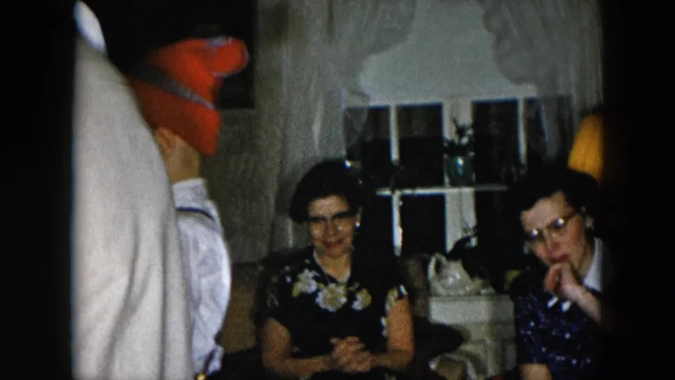 Two women in a 1950s domestic interior; woman in dark floral dress with glasses at center, second older woman in lighter clothing at right, partial figure at left frame edge.